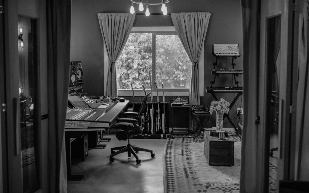 Cozy music studio with mixing console, guitars, and synthesizers near window with curtain, black and white.